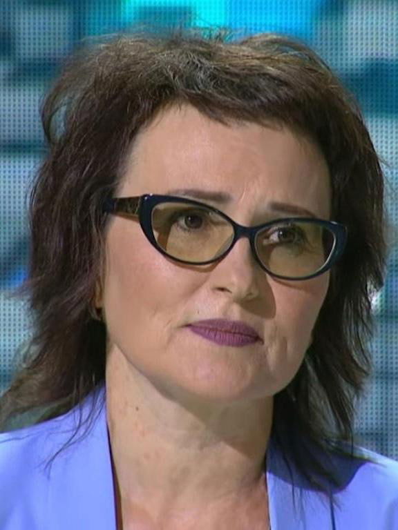 Крупный план другой женщины в очках во время интервью: короткие тёмные волосы, серьёзное выражение и студийный фон.