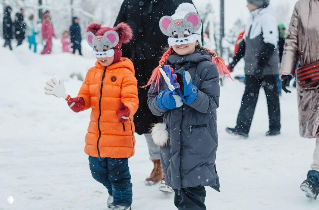Дети в зимней одежде и масках идут по праздничной площади в снегопад, создавая семейную и детскую атмосферу масленичных праздников.