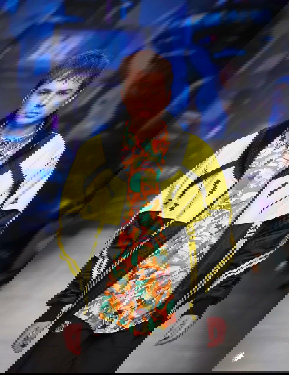 Мужская модель в жёлто‑чёрной куртке и яркой рубашке на фоне цифрового бэкграунда подиума — урбанистическая интерпретация народной эстетики.