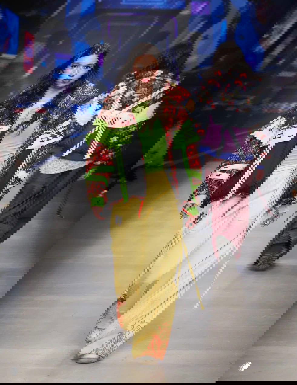 Модель в тёмно‑красно‑зелёной куртке с неоновыми акцентами и желтыми брюками на подиуме, где цифровые паттерны перекликаются со славянскими орнаментами.