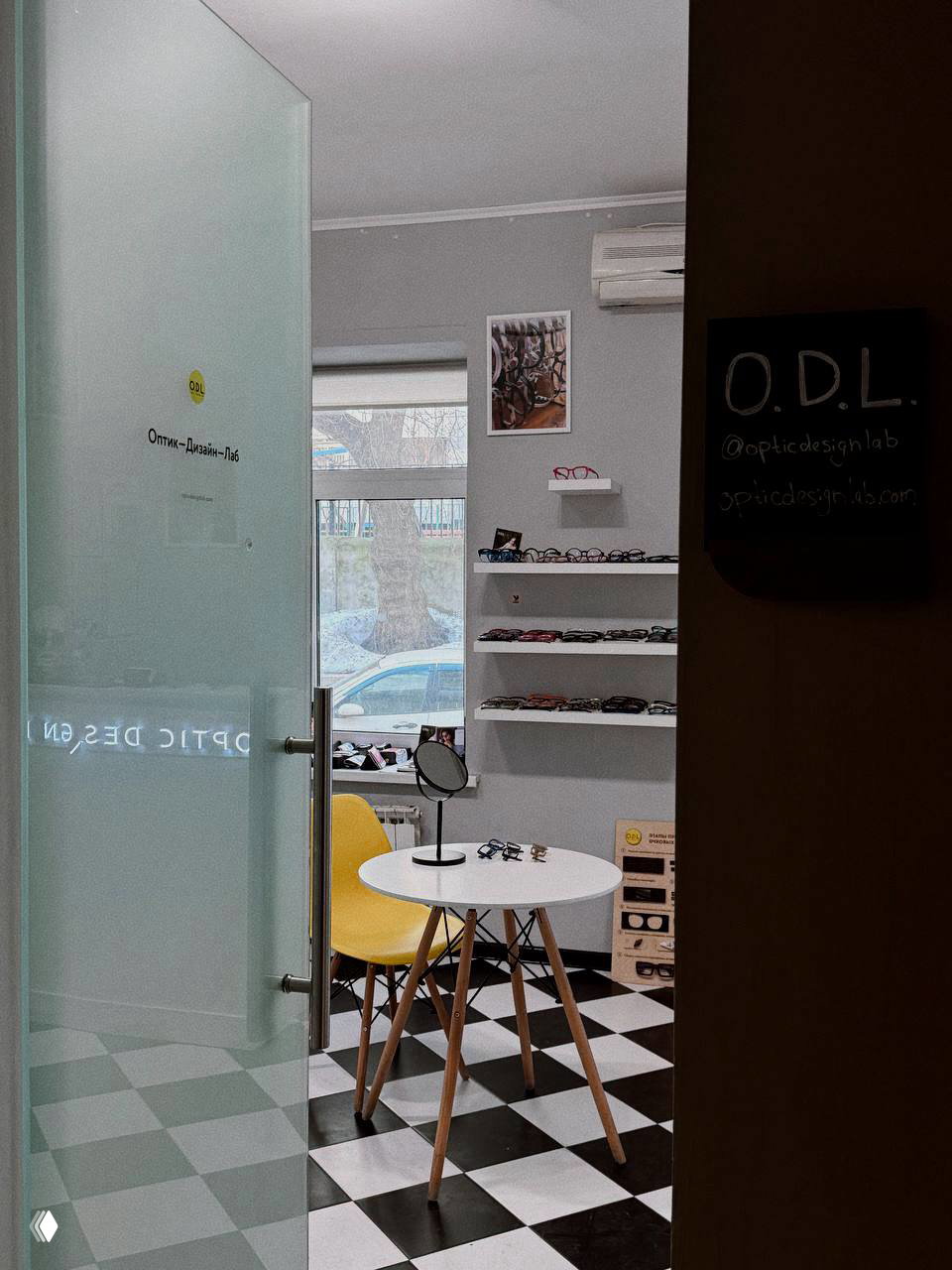 Интерьер шоурума O.D.L.: открытая стеклянная дверь, небольшой стол, жёлтое кресло и стеллажи с подборкой дизайнерских оправ на фоне помещений.