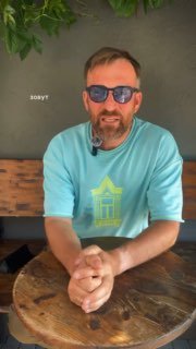 Мужчина в тёмных солнцезащитных очках O.D.L., в голубой футболке и с лёгкой щетиной, сидит за круглым деревянным столом на фоне тёмной стены.