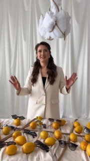Женщина в светлом пиджаке стоит за столом с лимонами и несколькими солнцезащитными очками; белые шторы создают минималистичный фон.