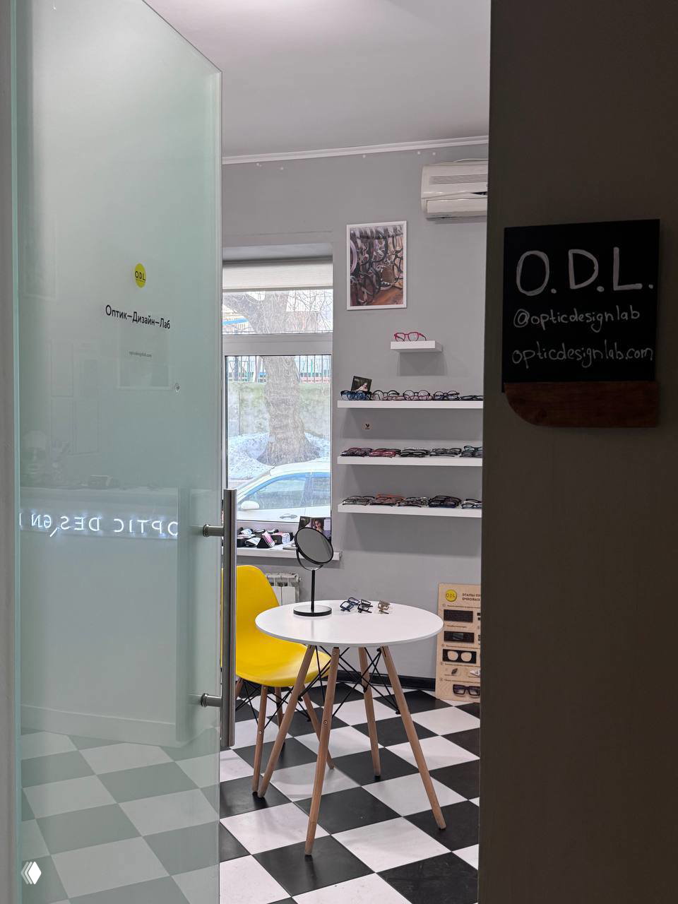 Вход в шоурум O.D.L.: стеклянная дверь с логотипом, внутри белый стол и жёлтый стул, стенды с оправами и небольшая табличка с надписью O.D.L.