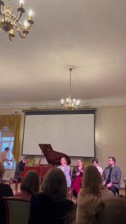 Сцена с пианино и сидящими выступающими — панель спикеров на дебатах по психоанализу и гештальту, видна часть аудитории.