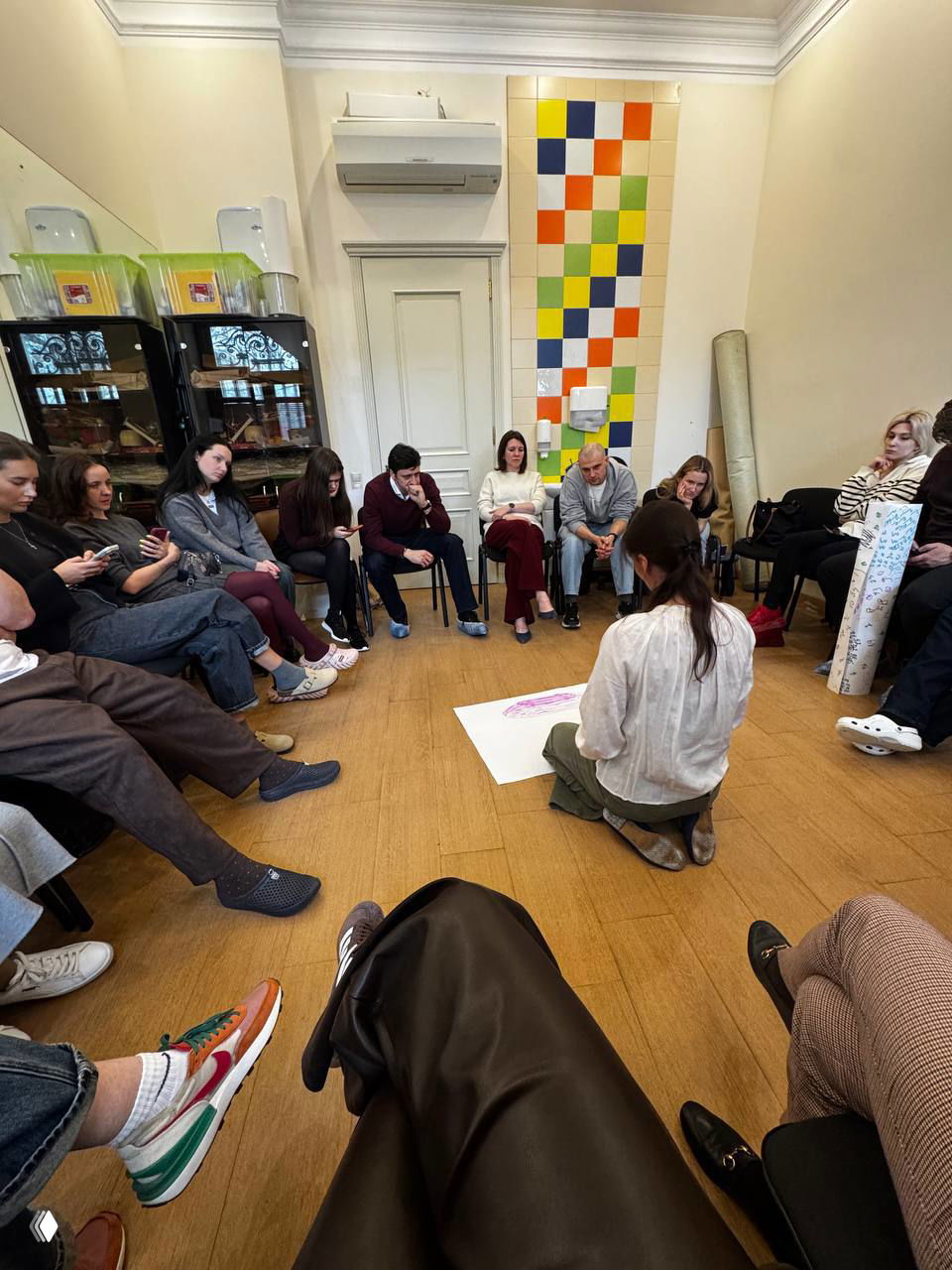 Круг участников в терапевтическом помещении: женщина на коленях рисует на листе на полу, вокруг сидят коллеги и внимательно наблюдают за процессом.