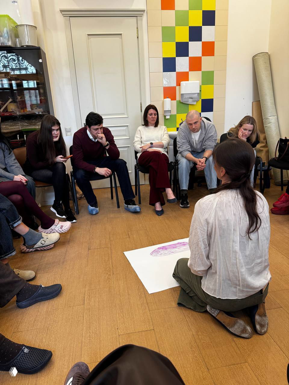 Вид сзади на участницу, сидящую на коленях и рисующую на листе; на заднем плане коллеги в креслах и заметная цветная плитка на стене кабинета.