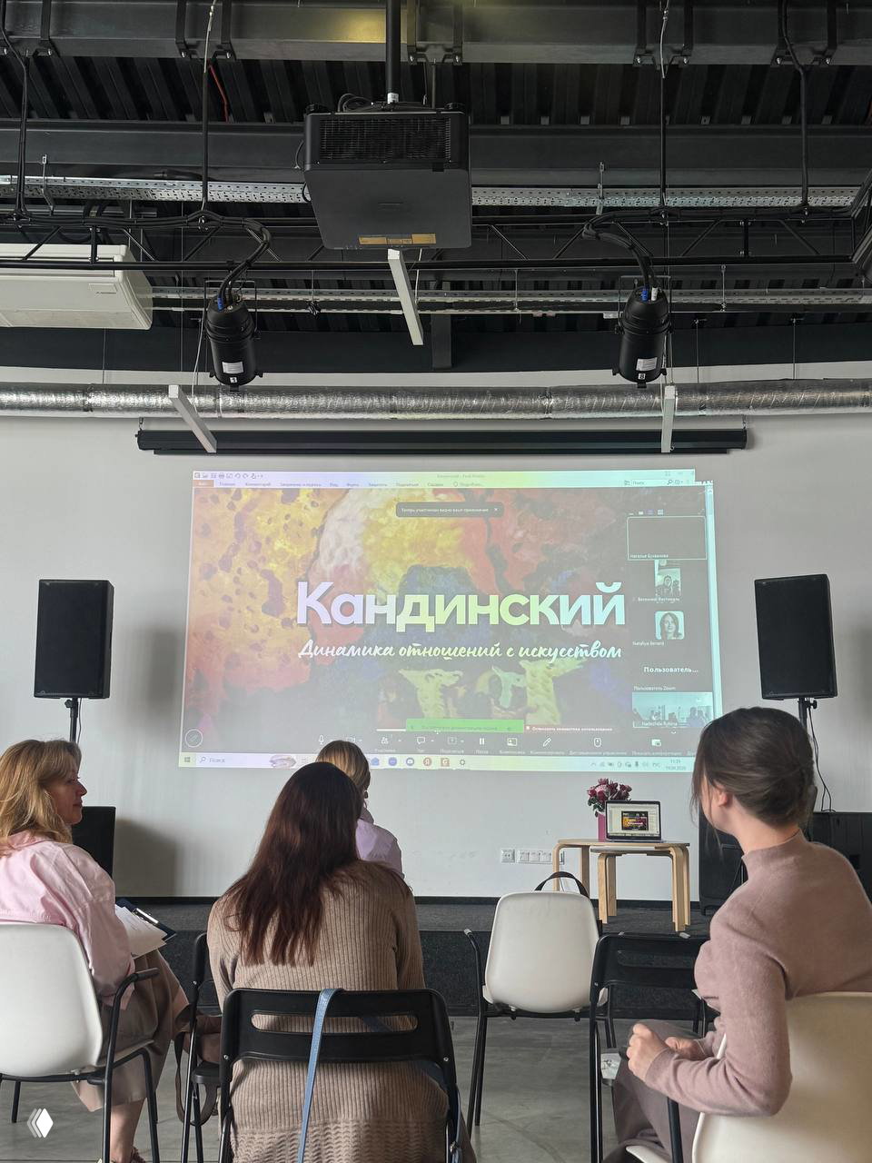 Аудитория смотрит на проекционный экран с презентацией «Кандинский — Динамика отношений и искусство», участники в сидячем зале наблюдают за докладом.