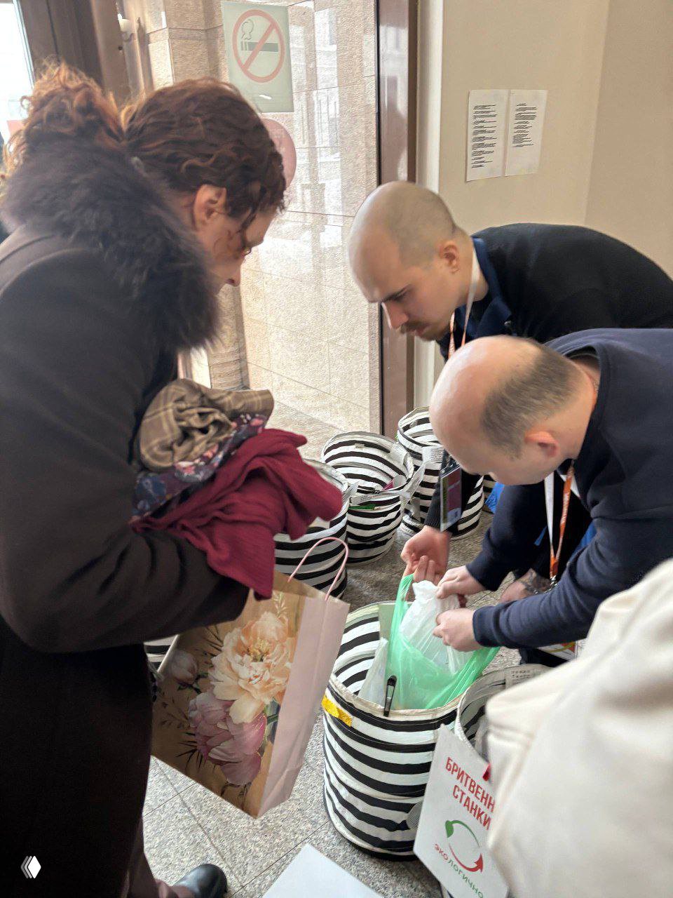 Волонтёры внимательно сортируют принесённые вещи: мужчина в бейдже и люди рядом кладут пакеты в соответствующие мешки для переработки.