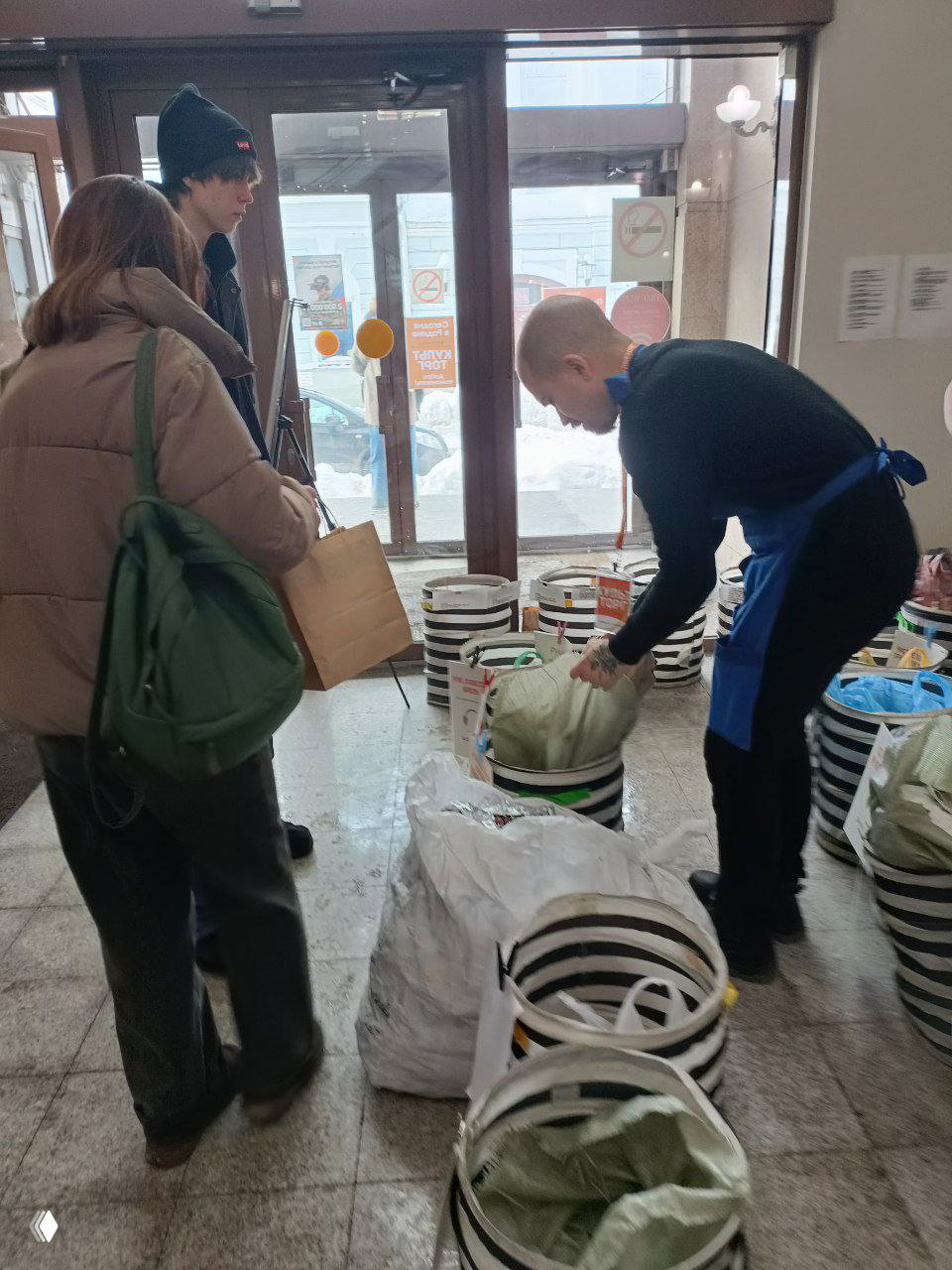 Волонтёр в фартуке помогает посетителю опорожнить пакет в корзину; видна организация приёма и подписи на мешках для разных материалов.