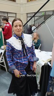 Елена, мастерица из Йошкар-Олы, сидит за столом на КультТорге в клетчатой рубашке с белым воротником, рядом её изделия и стенд фестиваля.