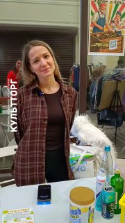 Улыбающаяся женщина за столом на фестивале КультТорг: образцы вторсырья — пластиковые и стеклянные бутылки, упаковка и принадлежности для сортировки отходов в помещении.