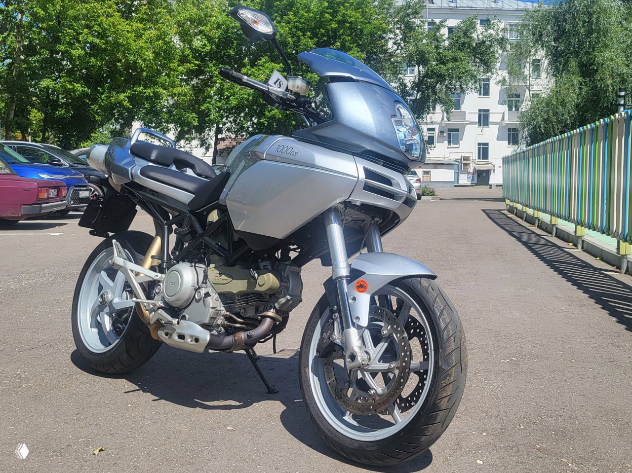 Трехчетвертной вид серебристой Ducati Multistrada 1000DS на стоянке: переднее крыло, рама и диски видны на фоне деревьев и зданий. Фото для объявления и оценки состояния.