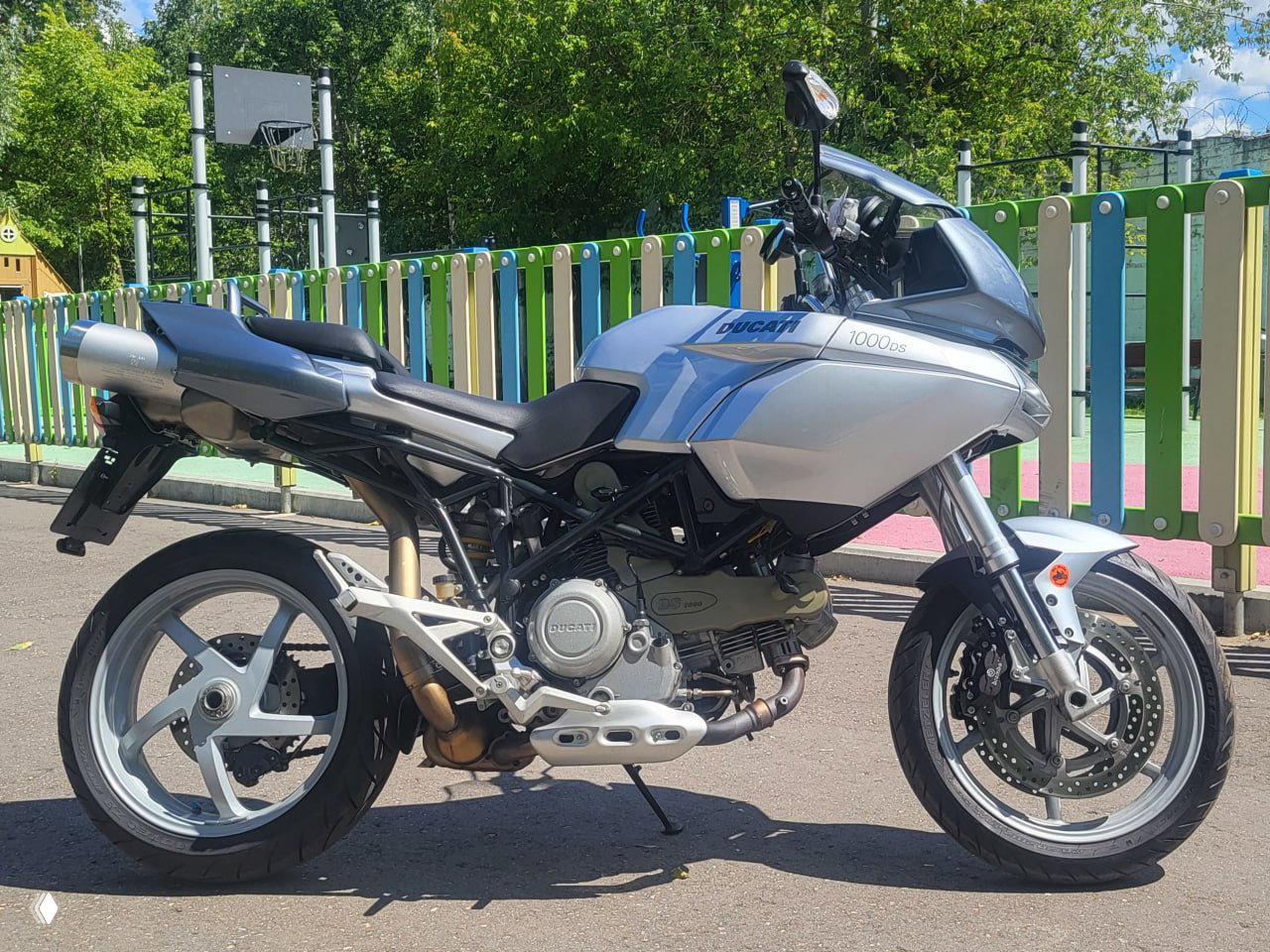 Боковой ракурс Ducati Multistrada 1000DS в серебристом цвете: бак, рама и выхлоп на асфальте, мотоцикл на боковой подножке в солнечный день. Фото для объявления и оценки состояния.