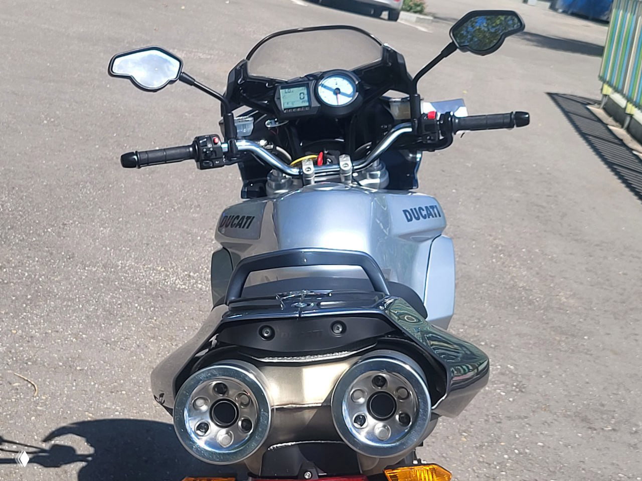 Вид сверху и сзади на приборную панель Ducati Multistrada 1000DS: руль, спидометр и двойной выхлоп на фоне городской среды. Фото для объявления и оценки состояния.