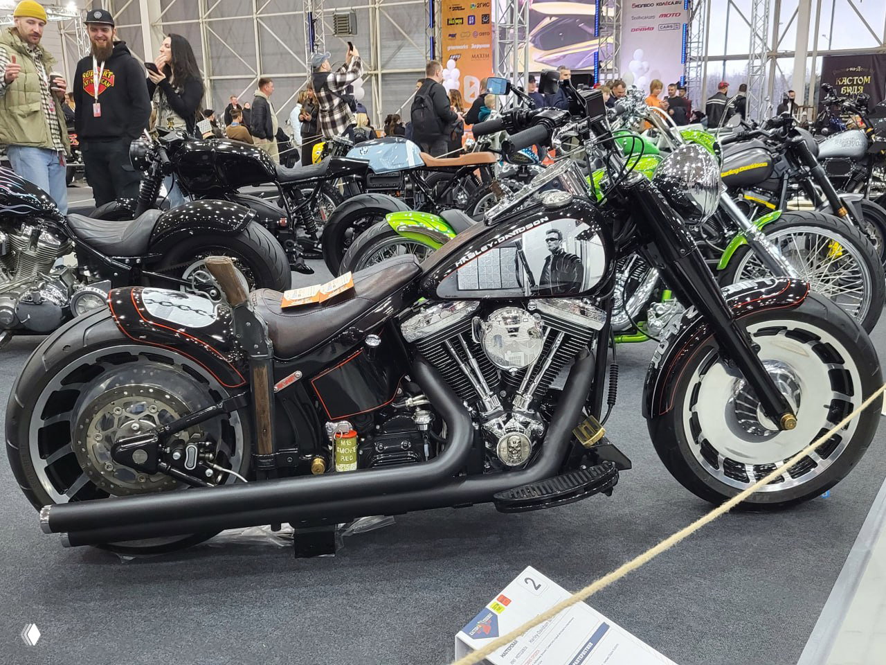 Чёрный кастомный бэггер на выставке: массивное заднее колесо, фрезерованные алюминиевые детали и толпа посетителей на заднем плане на стенде кастомов.