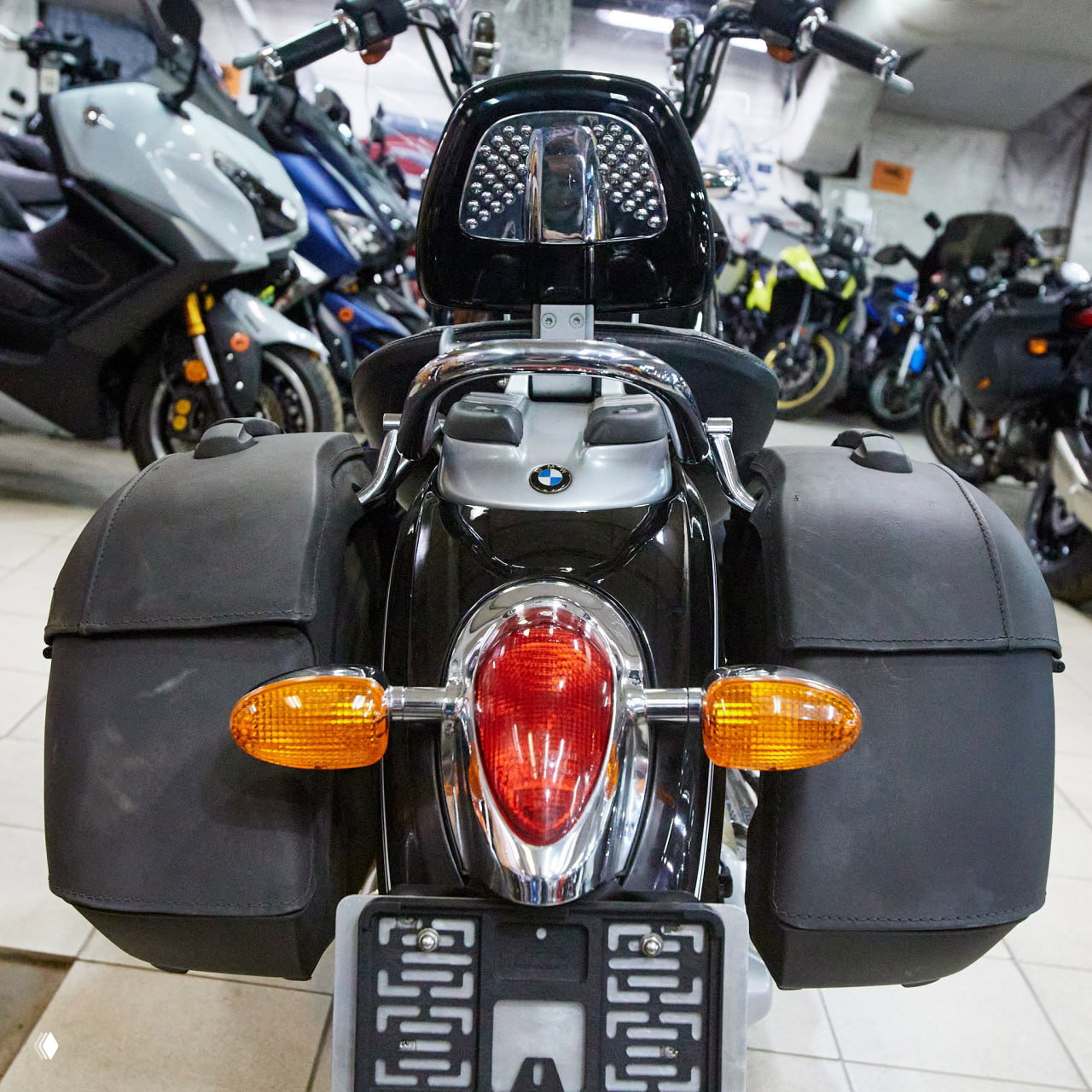Вид сзади на BMW R 1200 C: багажные сумки, задний фонарь и логотип BMW, мотоцикл стоит в ряду других моделей в шоуруме.