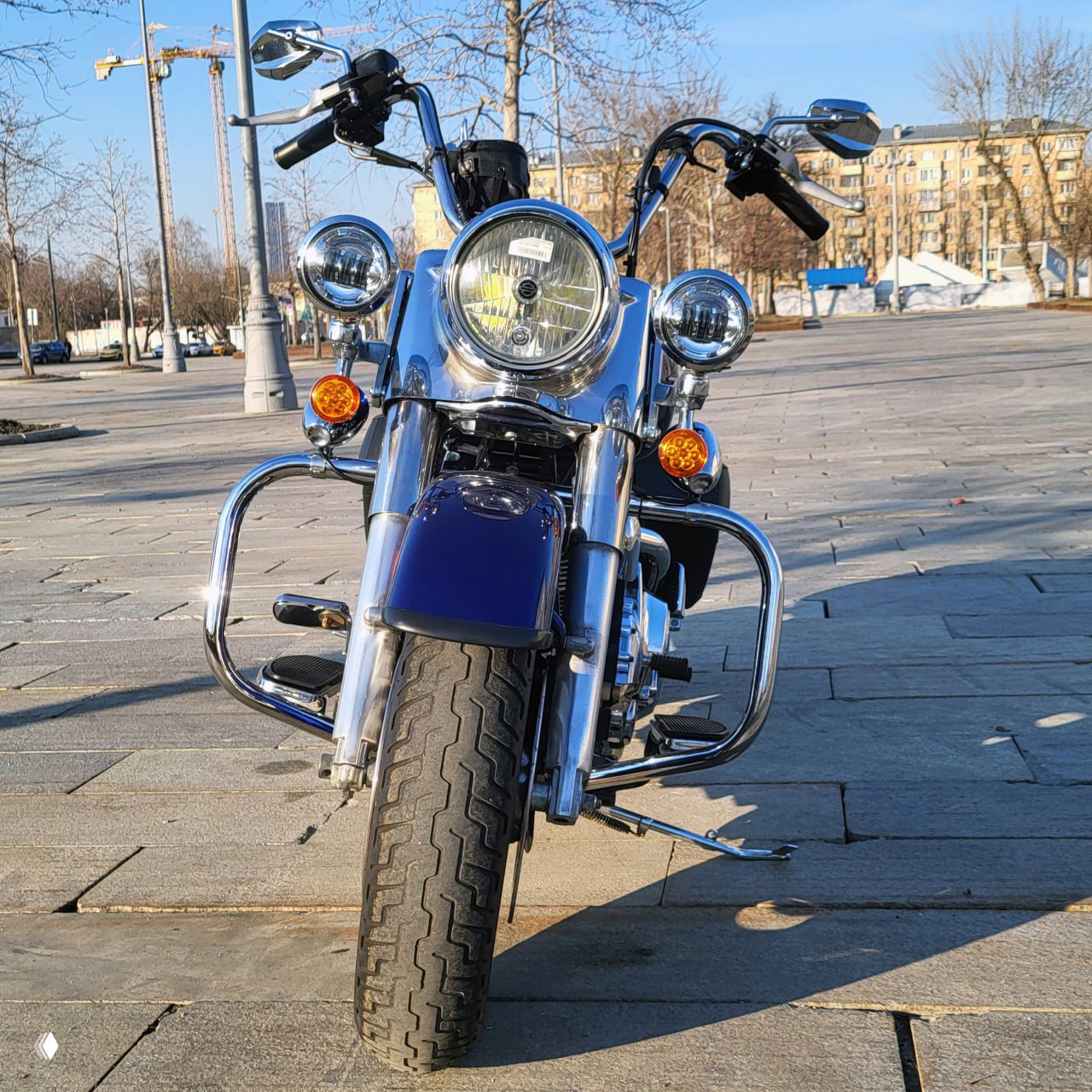 Фронтальный ракурс Harley Road King: крупный круглый фара, дополнительные фонари, хромированные дуги и широкая передняя покрышка на плитке площади.