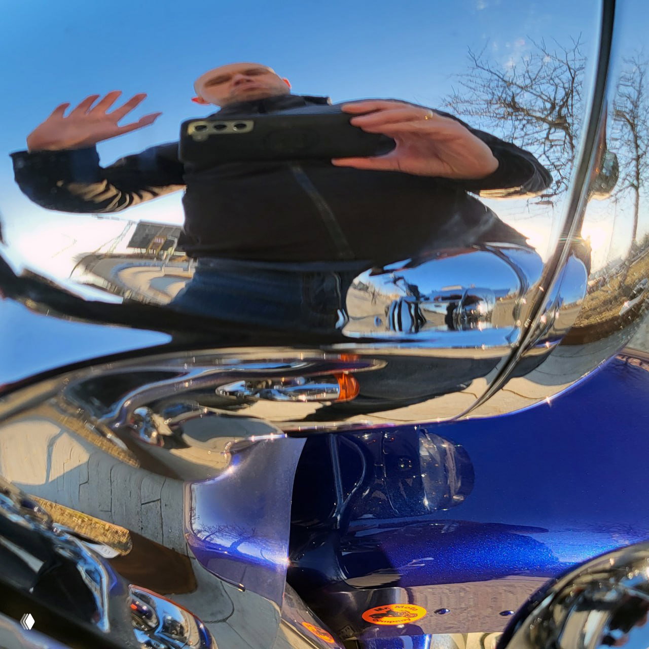 Отражение фотографа в хромированной крышке мотоцикла: видно корпус мотоцикла, отражённый городской пейзаж и сам автора снимка.