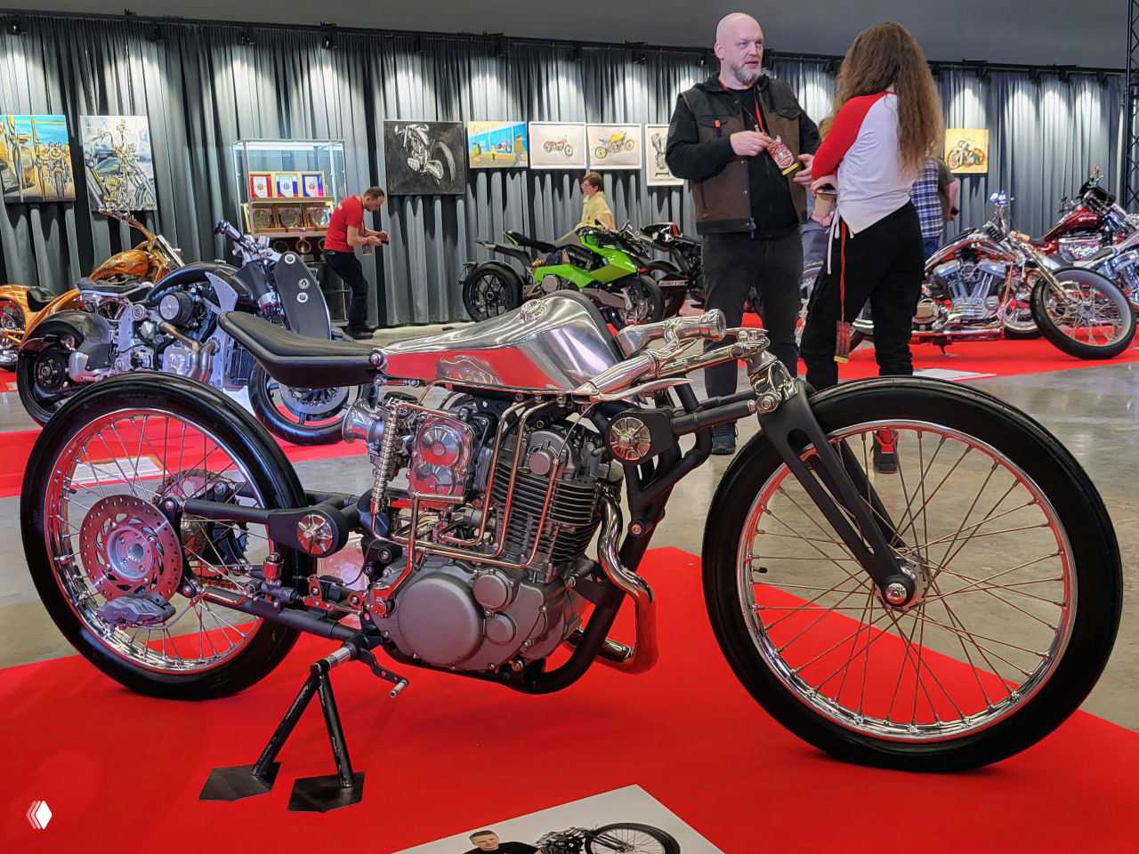 Тонкая хромированная рама классического кастома с минималистичным сиденьем и большим передним колесом на красном покрытии в музее.