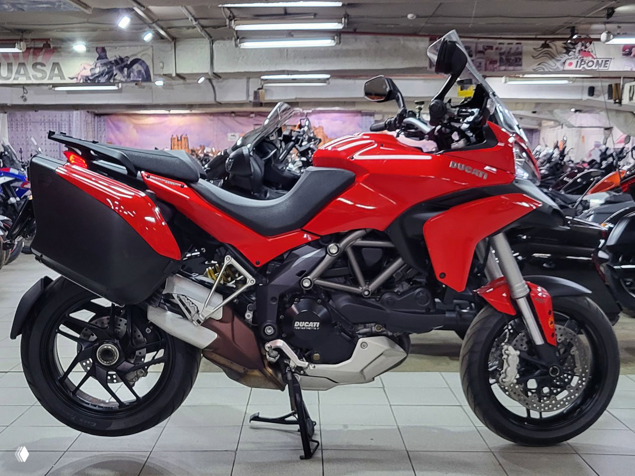 Боковой профиль красной Ducati Multistrada 1200 S в выставочном зале: рама, двигатель, багажные кофры и центральная подставка видны в кадре.
