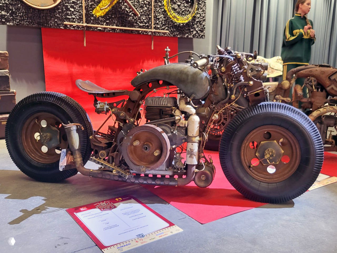 Низкий профиль кастомного мотоцикла с видимым двигателем и большим количеством хромированных элементов на фоне посетителей выставки.