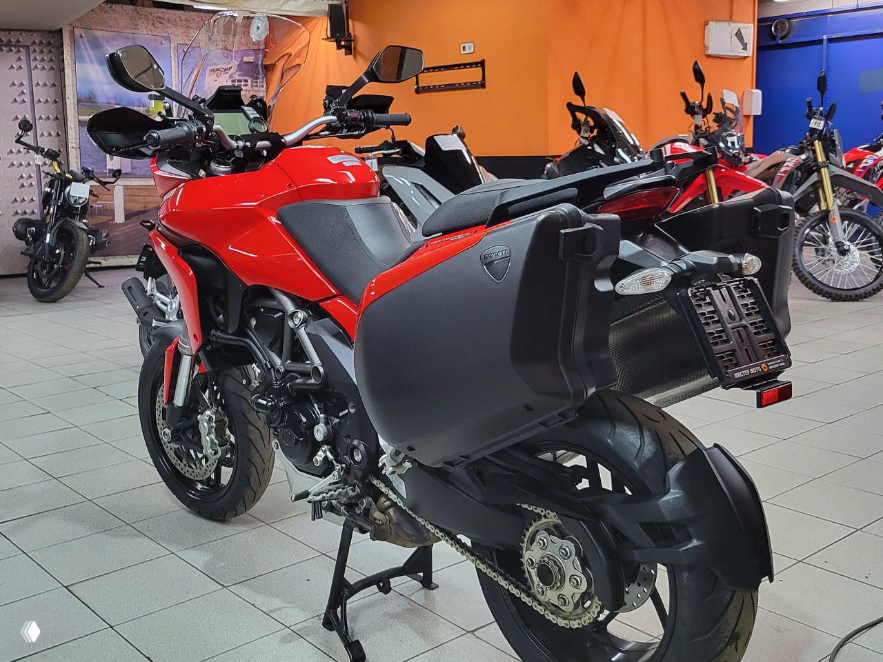 Задне-боковой ракурс Ducati Multistrada 1200 S с установленными боковыми кофрами, видны задняя часть, цепь и подножка, мотоцикл на подставке.