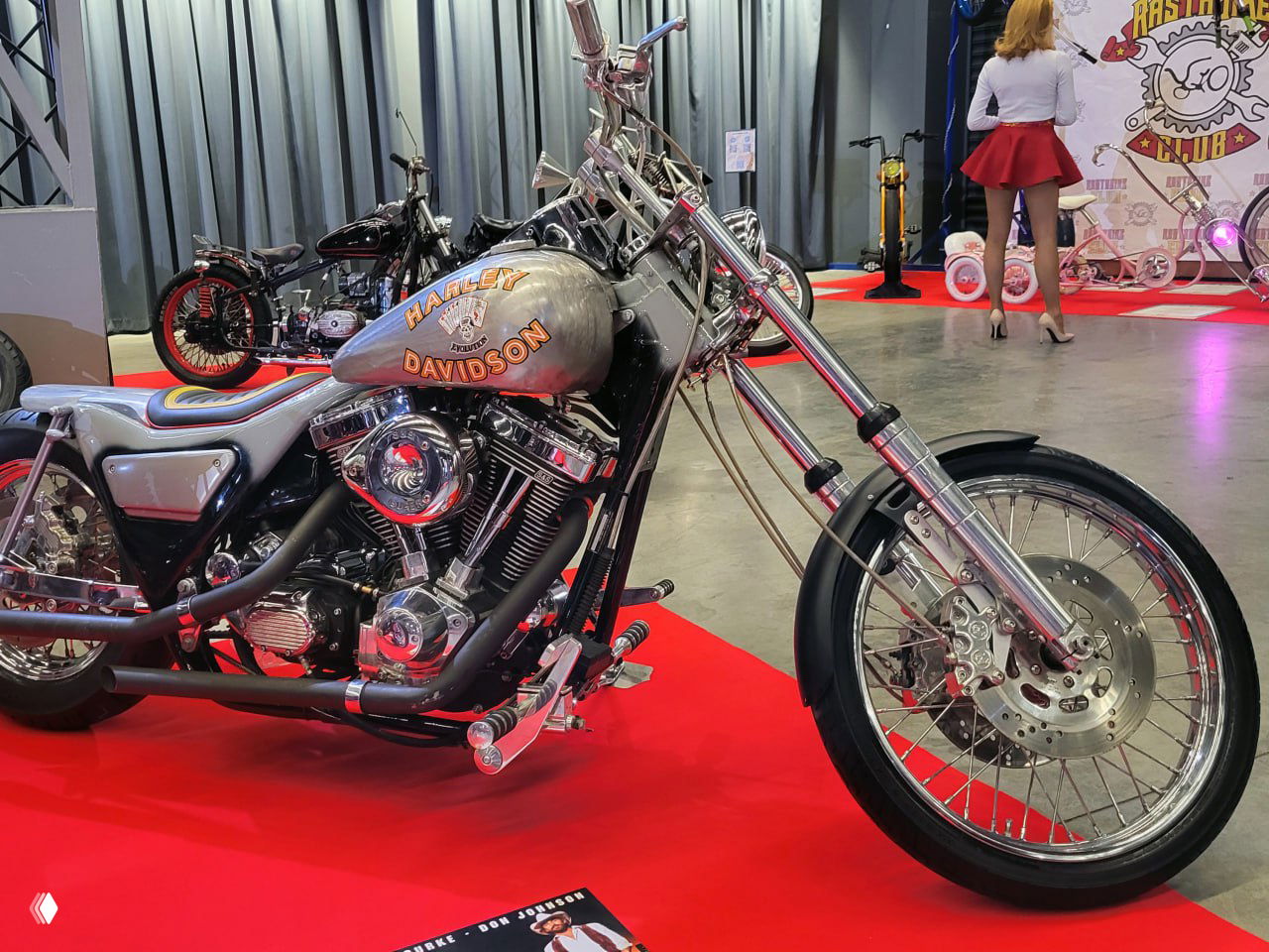 Необычный трёхколёсный или экспериментальный агрегат с массивными шинами и промышленным видом, выставленный на красном ковре среди других работ.