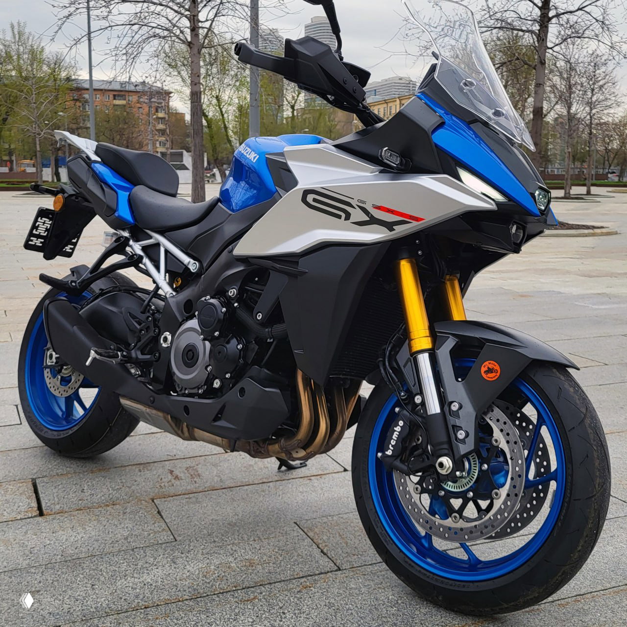 Передне-боковой ракурс синего Suzuki GSXS1000GX с золотыми вилками и суппортом Brembo на парковочной плоскости городского сквера.