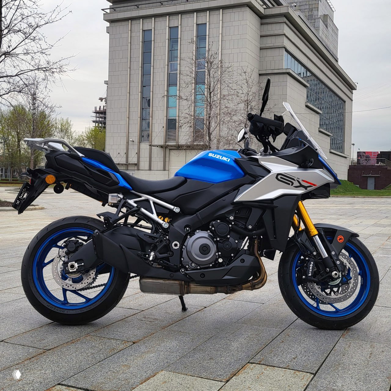 Боковой профиль Suzuki GSXS1000GX полностью в раме, мотоцикл на центральной части площади на фоне городских зданий, видна выхлопная система.