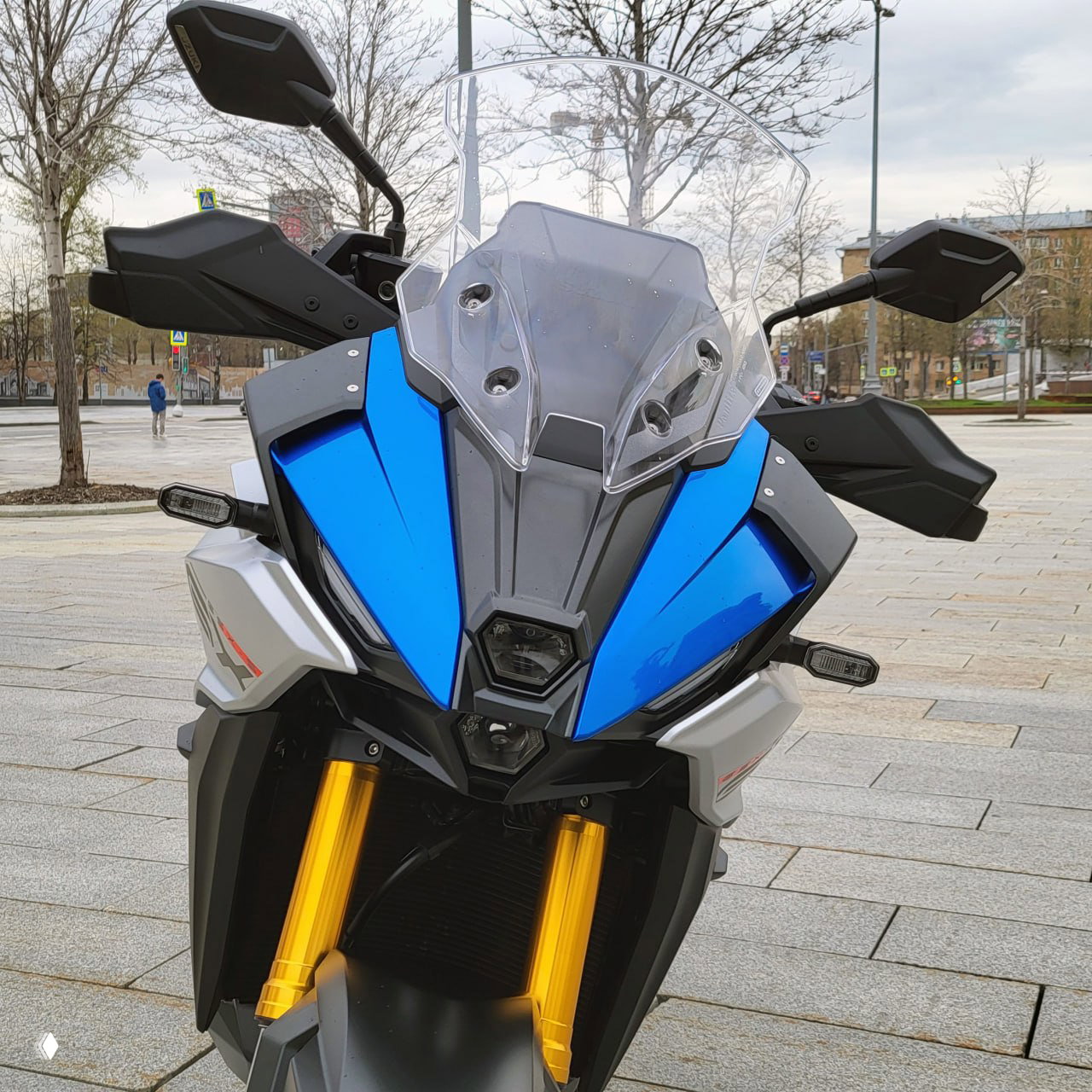 Фронтальная часть Suzuki GSXS1000GX с высоким ветровым стеклом и пластиковыми защитами рук, крупный план на облицовку и фары.