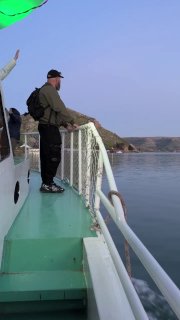 Человек стоит на носу лодки и смотрит на широкую реку и берег; кадр экспедиции на Евфрате, спокойная вода и прибрежный ландшафт.