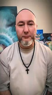 Портрет мужчины в светлой футболке с четками и крестом на шее, на фоне абстрактных картин; кадр из видео.