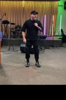 Мужчина с микрофоном стоит на сцене в помещении для богослужений; на заднем плане музыкальное оборудование, световые полосы и занавеска.