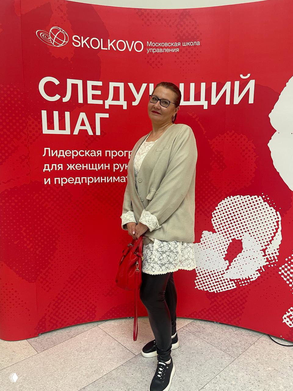 Женщина позирует на фоне яркого красного баннера Skolkovo с надписью «Следующий шаг»; фото с лидерской программы для женщин и предпринимательниц.