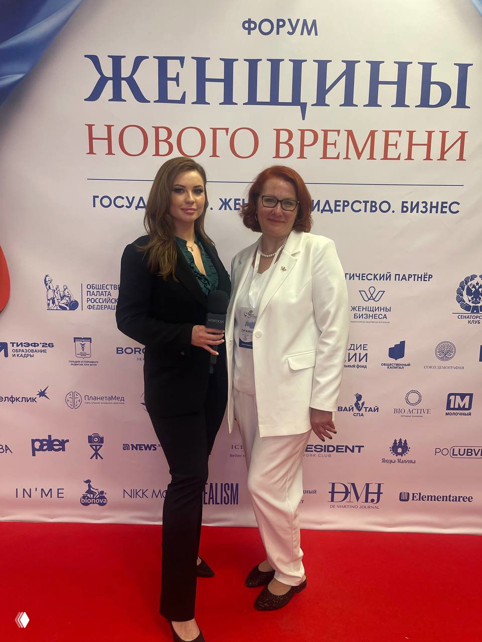 Две женщины позируют на красной дорожке у баннера форума «Женщины нового времени», микрофон и логотипы партнёров видны на фоне