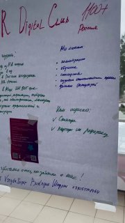 Фотография плаката с крупными рукописными заметками и прикреплённой печатной флайер-вкладышем, видна рабочая сессия фестиваля на фоне.
