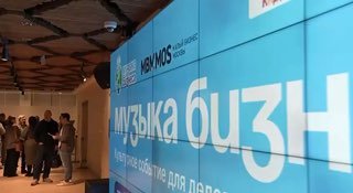 Широкий экран с надписью «Музыка Бизнеса» в фойе мероприятия, рядом люди и участники в коридоре перед началом события в Москве, атмосфера подготовки.