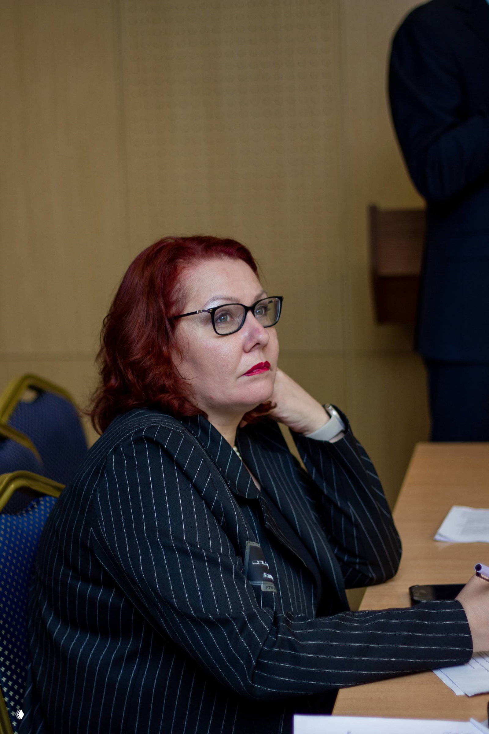 Крупный план участницы круглого стола: женщина в очках с рыжими волосами внимательно слушает доклад и делает пометки, атмосфера деловой дискуссии.
