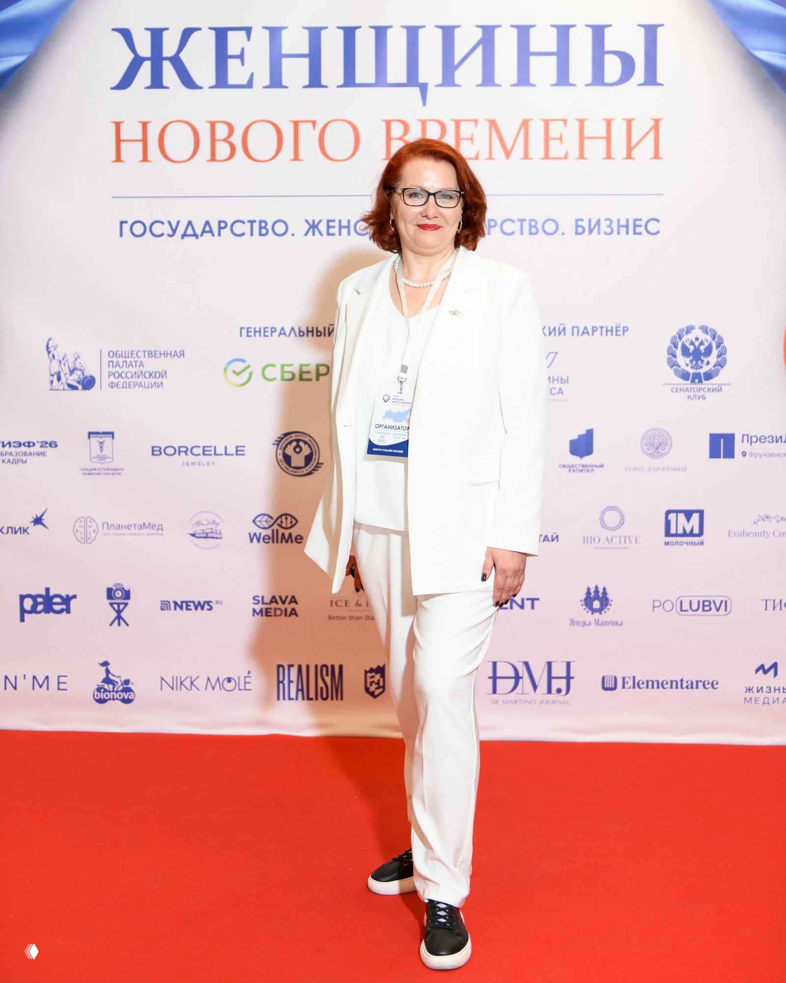 Женщина в белом костюме на красной дорожке перед баннером «ЖЕНЩИНЫ НОВОГО ВРЕМЕНИ», на фоне логотипы партнёров.