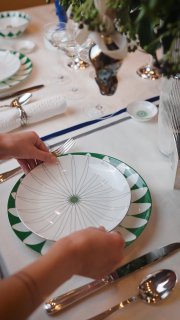 Руки ставят белую тарелку с зелёной каймой и геометрическим узором на праздничный стол с приборами, бокалами и зелёными декоративными ветками