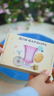 Руки держат подарочную карточку «Дом Фарфора» с рисунком чашки и элементов декора; на заднем плане видна синяя тарелка и размытая сервировка стола, контекст подарка.