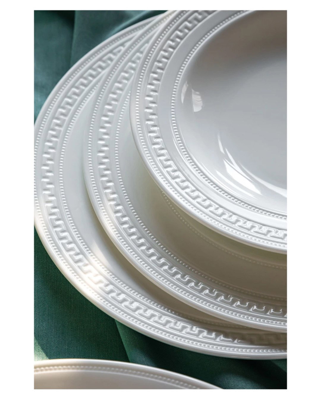 Крупный план кромки тарелок Wedgwood «Инталия»: рельефный меандр и жемчужные точки на белом костяном фарфоре, виден детализированный орнамент.