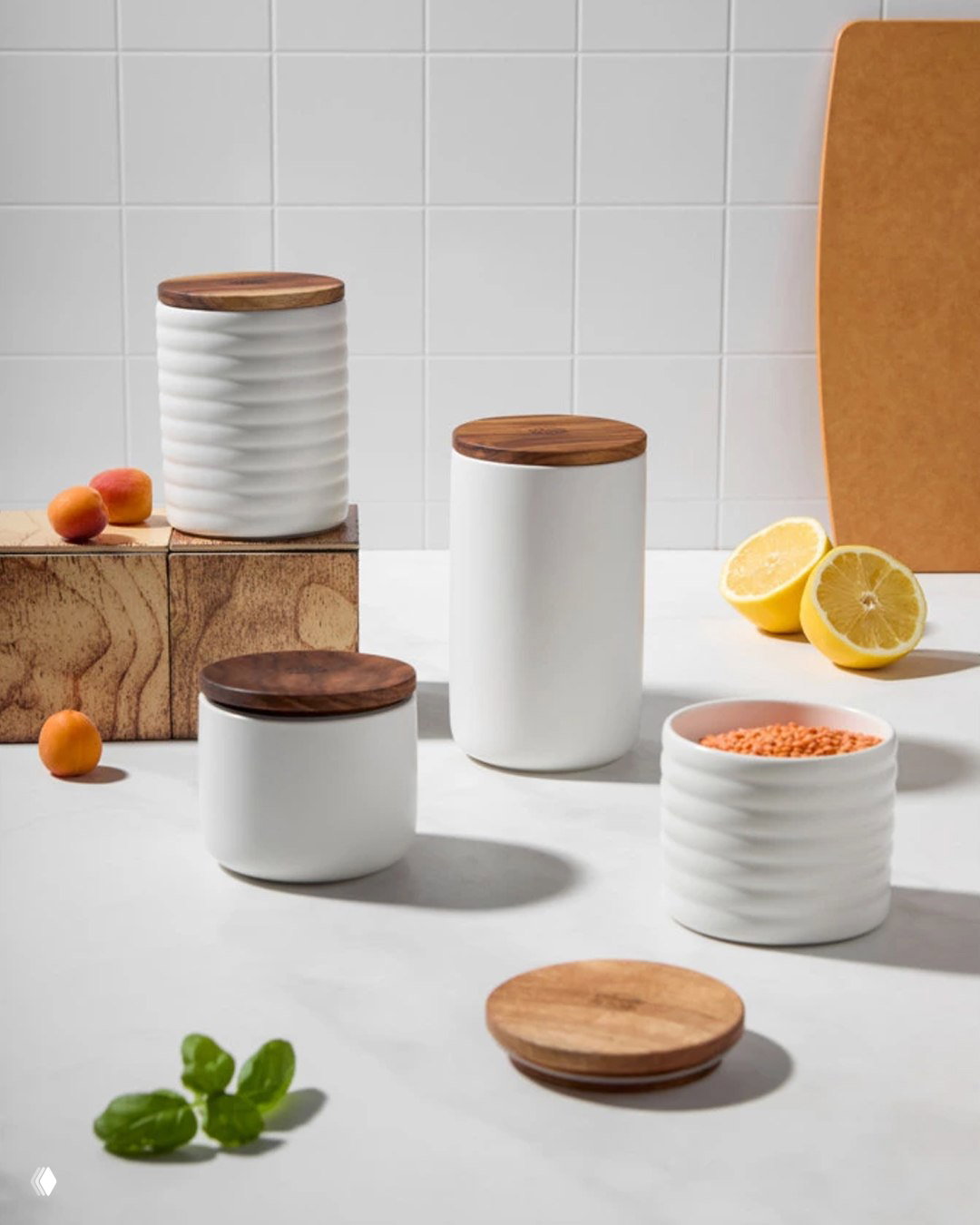 Белые керамические банки с деревянными крышками коллекции Decor de Table на светлой рабочей поверхности, стилизованная композиция с фруктами и специями.