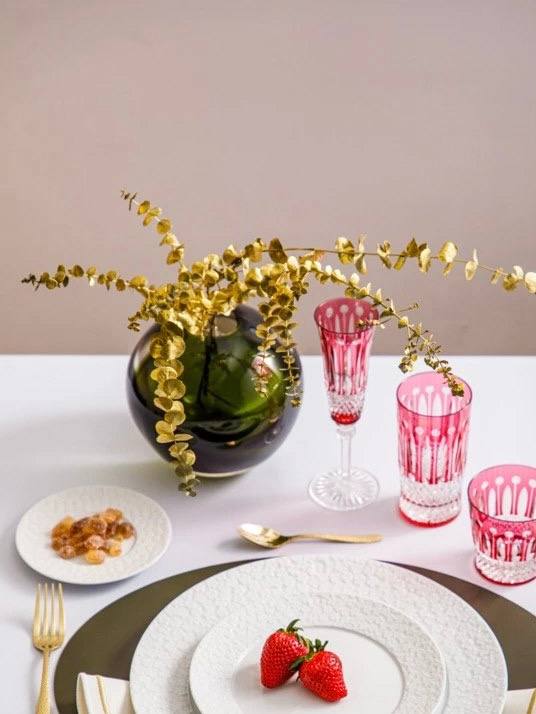 Сервировка с белыми тарелками, розовыми бокалами и маленькой круглой вазой с золотыми веточками — стильная композиция для стола.