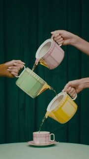 Три пастельных фарфоровых предмета Signum (лазурный, розовый, жёлтый) с ручной золотой окантовкой, из которых льётся чай на тёмно‑зелёном фоне