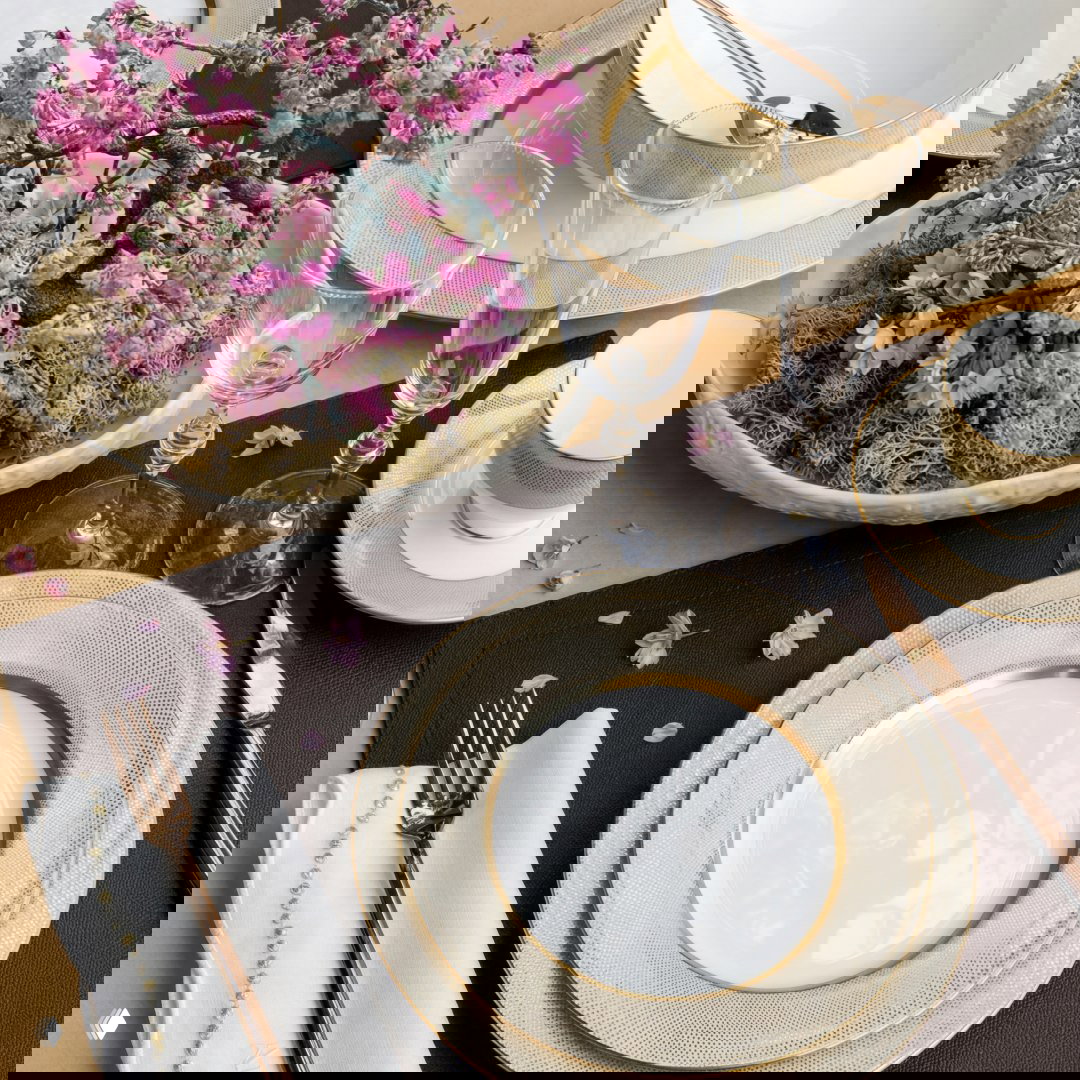 Стол с сервировкой: фарфоровые тарелки с золотой каймой, бокалы и декоративная композиция с розовыми цветами в центре, крупный план.