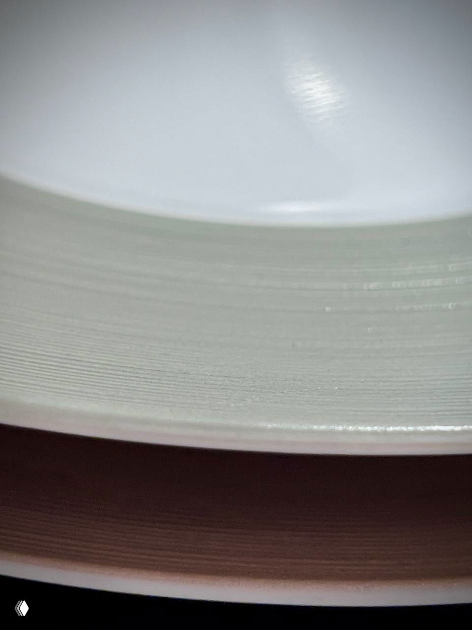 Край и бортик тарелки крупным планом: слоистая структура эмали, параллельные борозды и едва заметный зеркальный бликующий эффект на белом фоне.