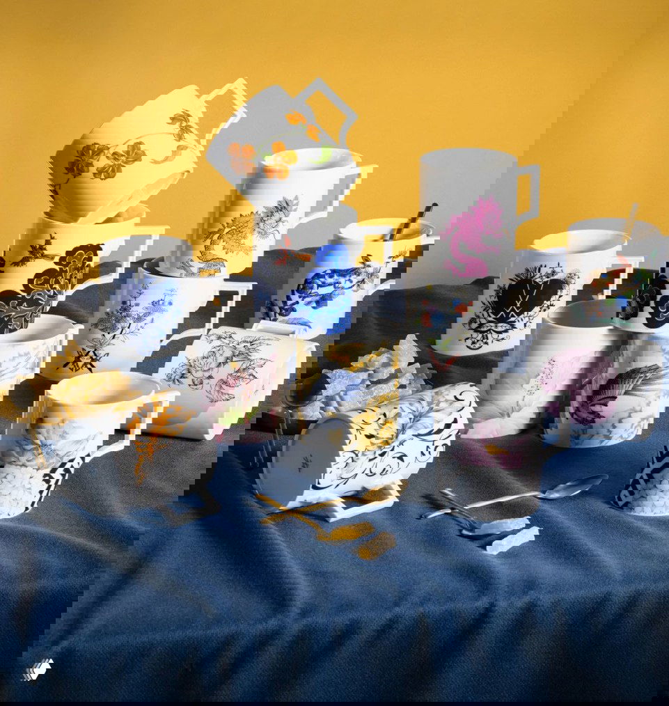 Коллекция декоративных фарфоровых кружек с растительными и орнаментальными рисунками, расположенные на ткани рядом с золотыми ложками.