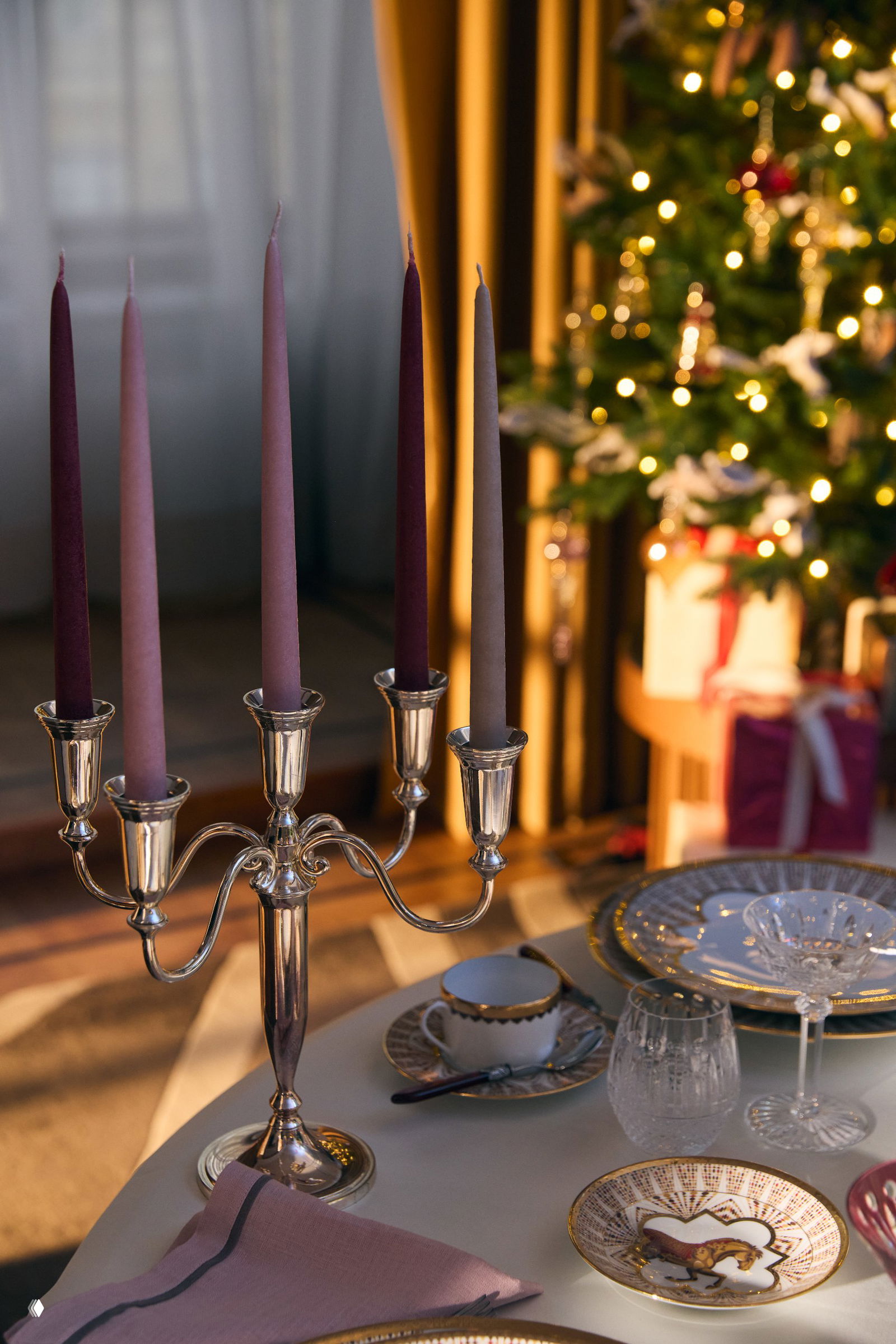 Крупный план серебряного канделябра с тонкими разноцветными свечами на фоне размытых огней новогодней ёлки.